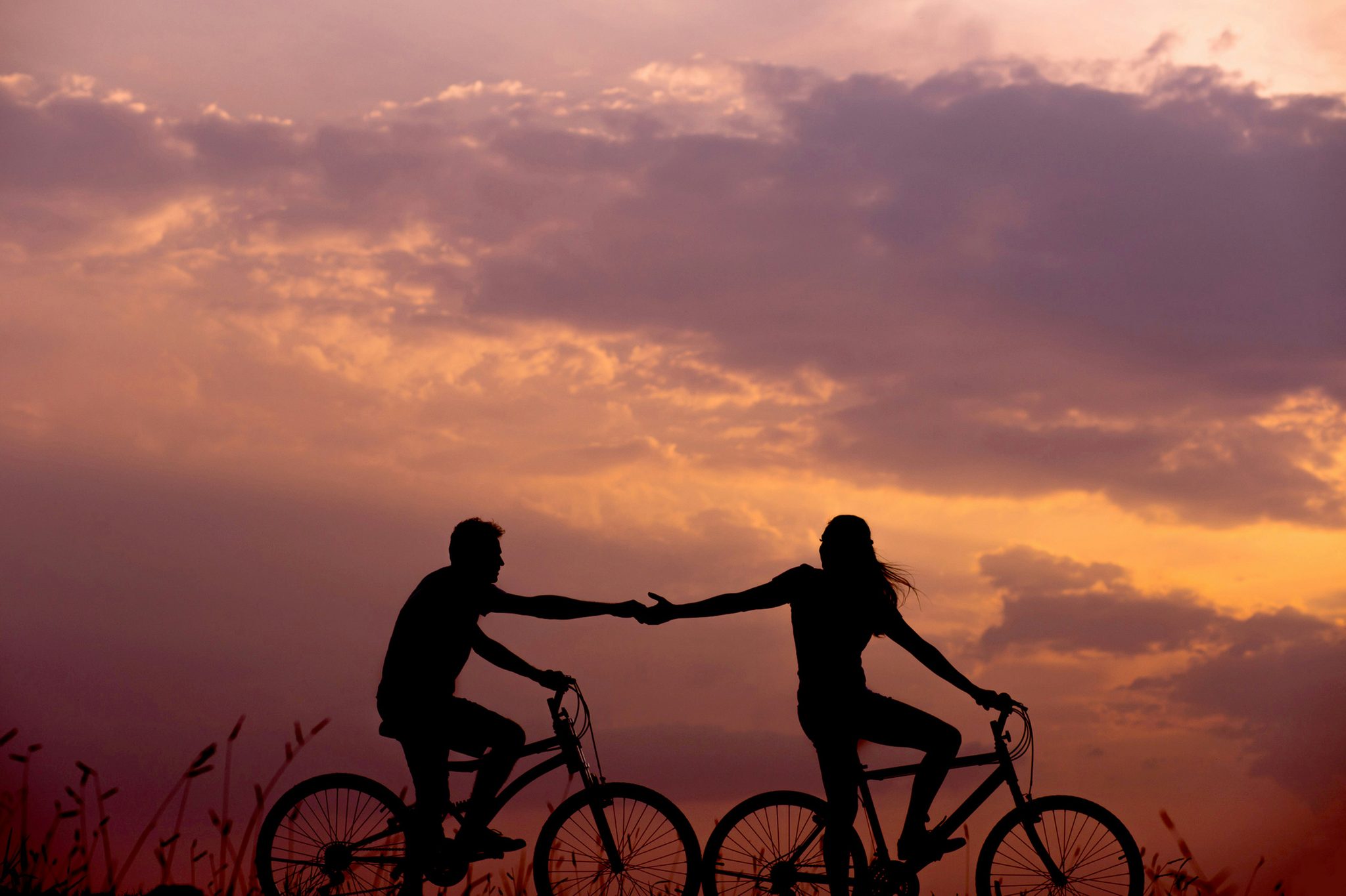 Two cyclists riding side by side at sunset, holding hands